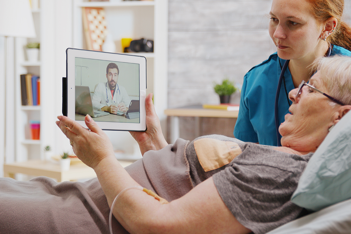 Old disabled woman lying in hospital bed having an online video call with a doctor