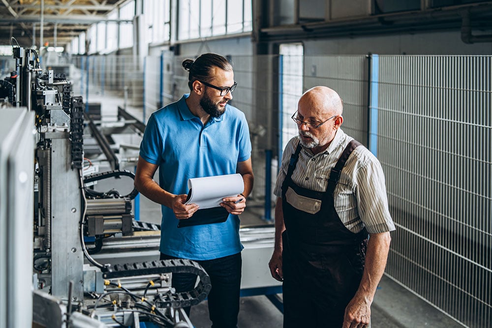 Young manager with beard showing and inspecting working process of adult professional worker on the big factory.